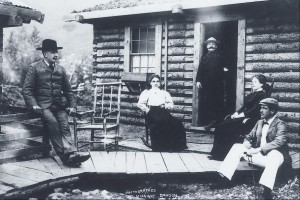 Des personnes assises jasent devant une maison en bois rond dans une photo datant du début du 20e siècle.