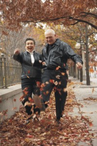Lors d'une marche automnale, un couple d'aînés, un homme et une femme, rient et balaient des feuilles mortes à grands coups de pieds.